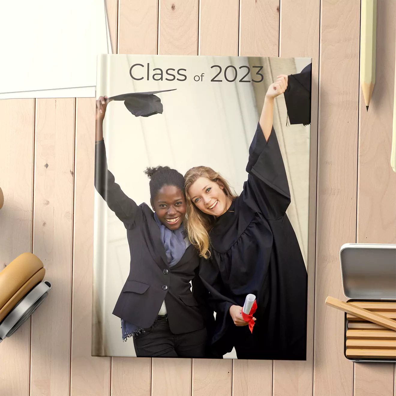 Photobook cover titled 'Class of 2023' featuring two graduates in caps and gowns celebrating. One graduate holds a diploma and the other raises a cap. The photobook is placed on a wooden surface with stationery items around.