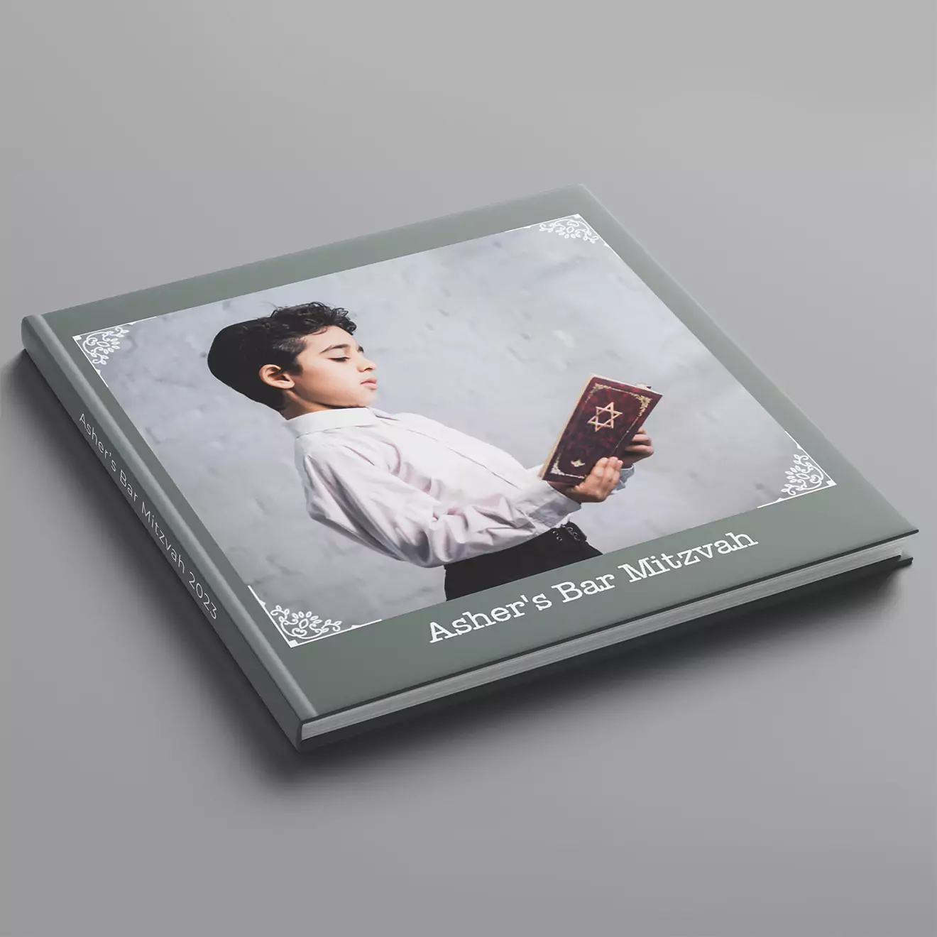 A custom photobook titled 'Asher's Bar Mitzvah' featuring a cover image of a young boy holding a book with a Star of David. The photobook has a grey cover with decorative white corners.