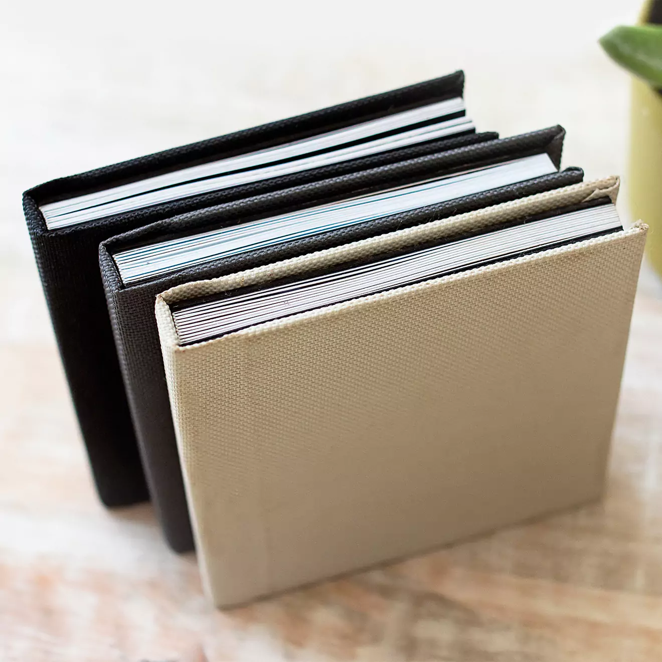 Three hardcover photobooks with textured covers in black and beige are standing upright on a light-colored surface. The photobooks are neatly aligned, showcasing their spines and pages.