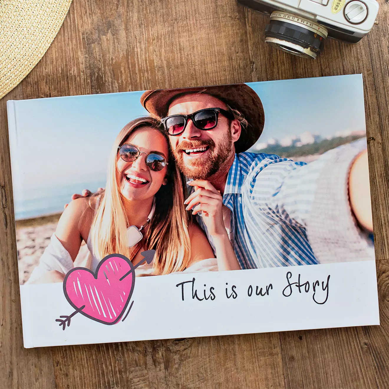 A photobook with a cover image of a smiling couple taking a selfie on a beach, featuring a pink heart with an arrow and the text 'This is our Story' on a wooden surface with a camera nearby.