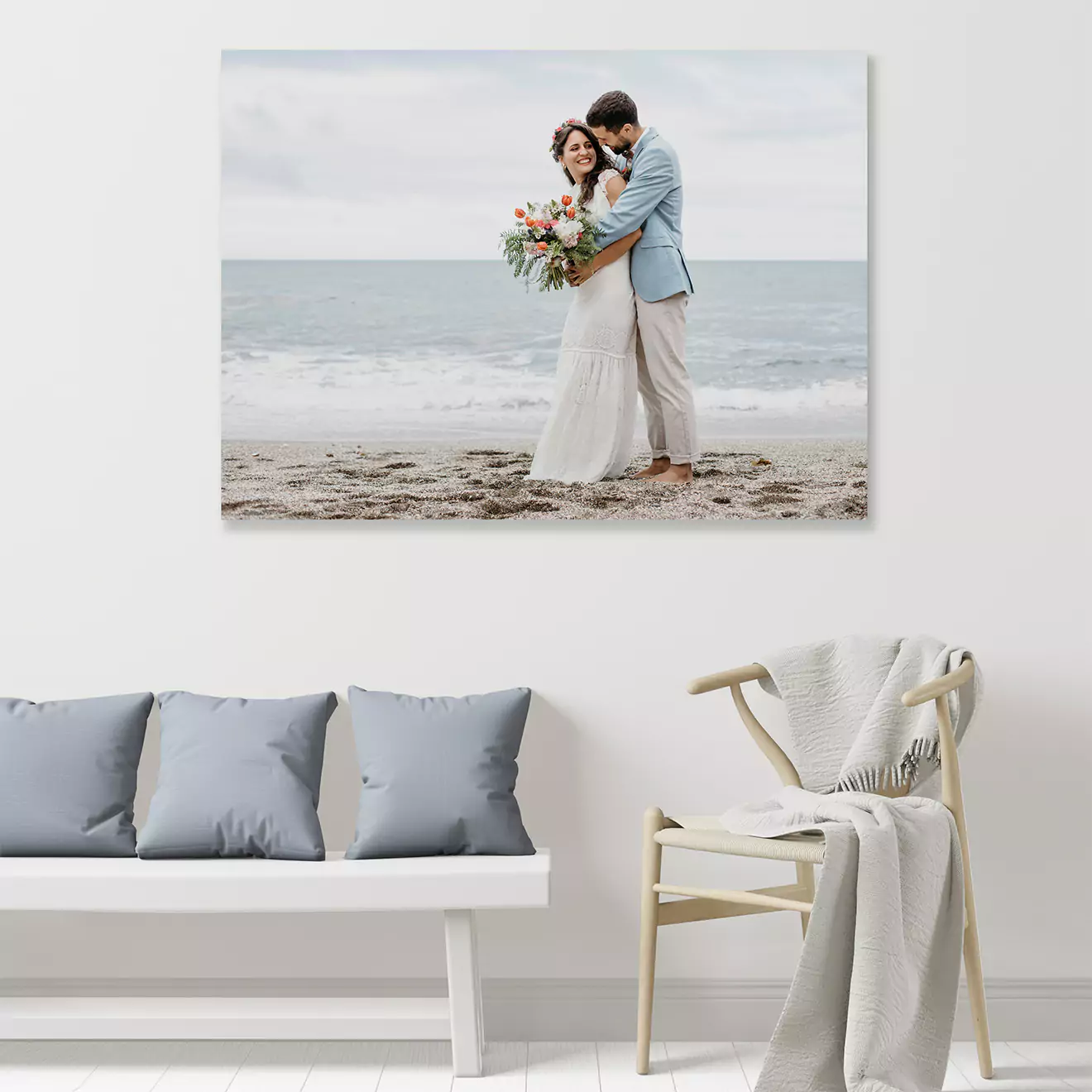 A stylish living room features a large wall art print of a couple embracing on a beach. The room includes a white bench with grey cushions and a wooden chair draped with a blanket.