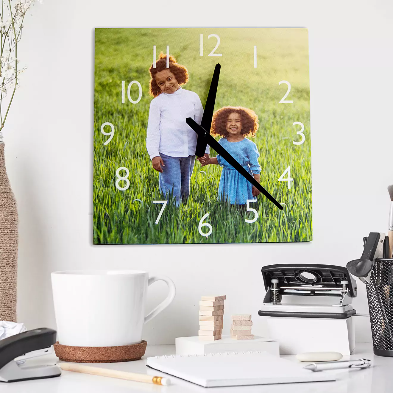 Square wall canvas clock featuring a photo of two children standing in a grassy field, with large white numerals and black clock hands. The clock is displayed above a desk with office supplies.