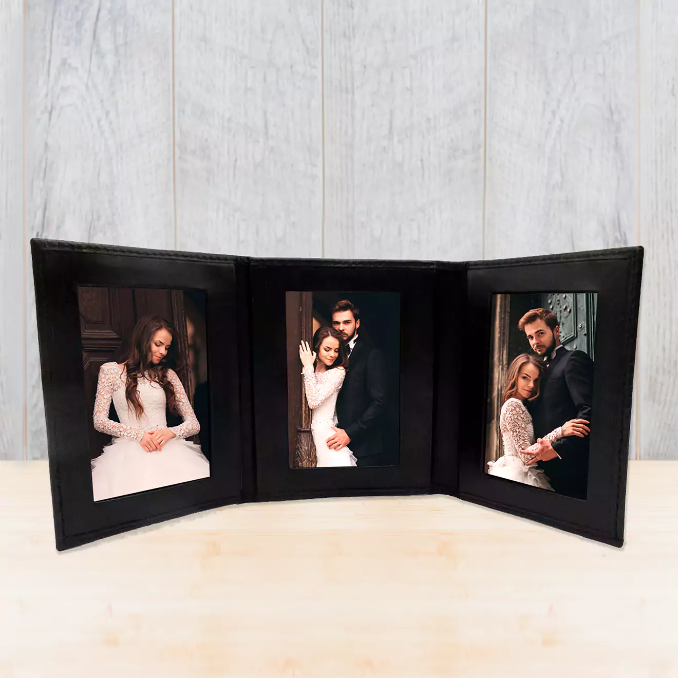 A black tri-fold photo frame displaying three wedding photos of a couple in elegant attire. The frame is set against a light wooden background, showcasing the couple's special moments.