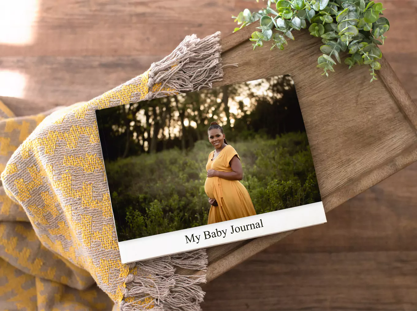 "Personalised photobook titled 'My Baby Journal' featuring a pregnant woman in a yellow dress standing in a lush green field. The photobook is placed on a wooden surface with a yellow and grey patterned blanket and a small potted plant nearby. Ideal for baby shower gift ideas and personalised gifts in South Africa."