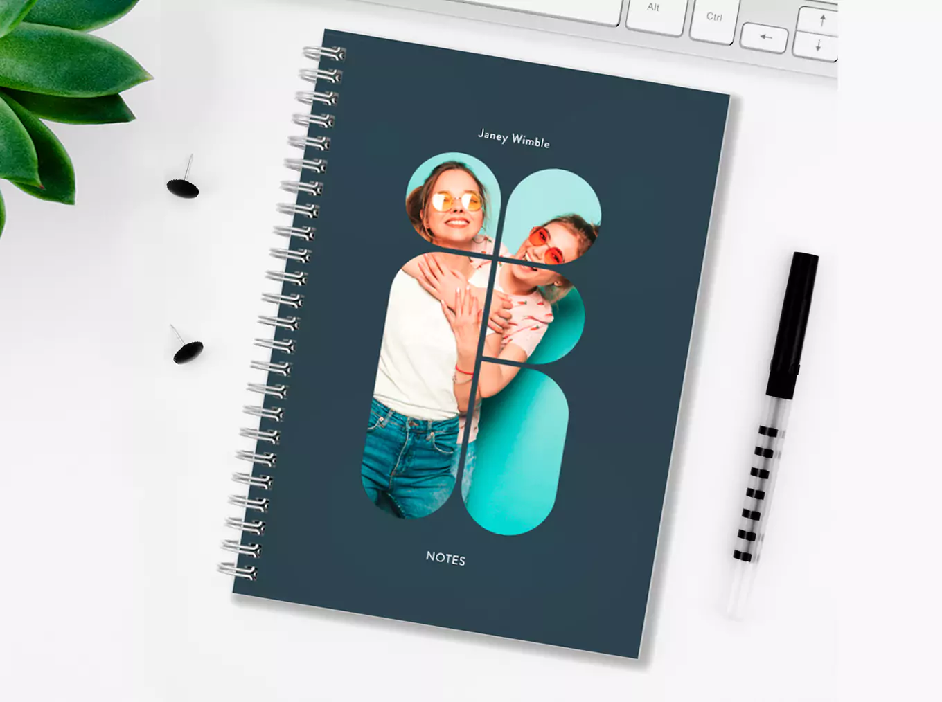 "Personalised spiral-bound notebook with a custom photo cover featuring two people, set against a dark blue background. The notebook is placed on a white desk alongside a black and white striped pen, a keyboard, and a green plant. Ideal for gifts and stationery lovers."