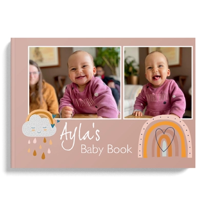 Hardcover baby photo book titled 'Ayla's Baby Book' featuring a pink cover with two photos of a smiling baby in a pink outfit. The design includes cute illustrations of a cloud with raindrops and a rainbow with a heart. Ideal for baby shower gift ideas and personalised gifts in South Africa.