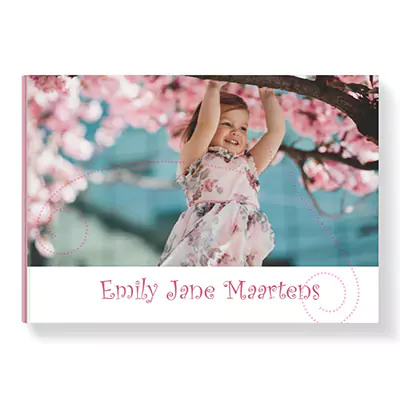 Personalised hardcover photo book with a 'Pink Curlz' theme, featuring a cover image of a young girl in a floral dress playing among cherry blossoms. The name 'Emily Jane Maartens' is printed in a decorative pink font on the white bottom border. Ideal for gifts such as baby showers, birthdays, or Christmas.