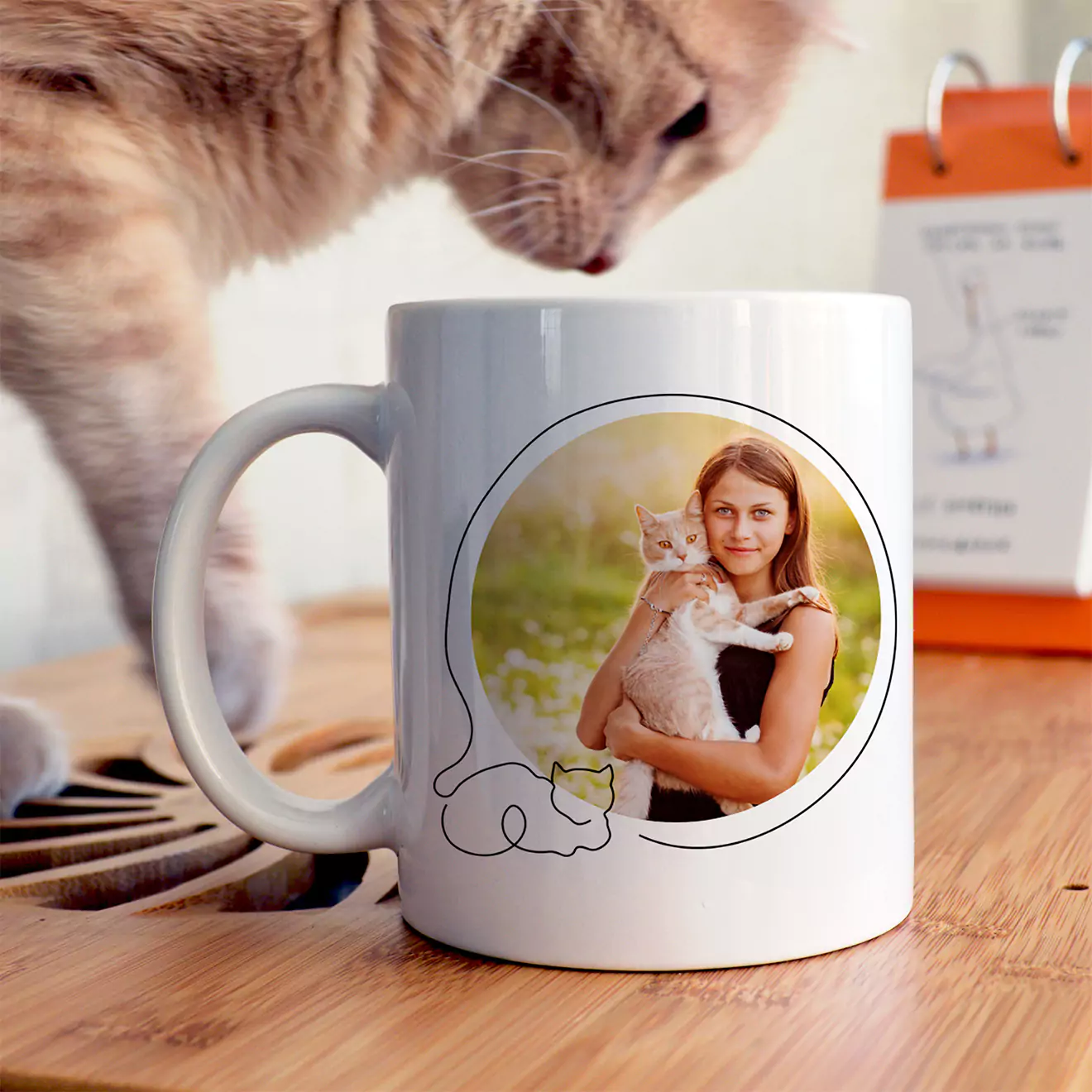 White ceramic mug featuring a personalised photo of a woman holding a cat, with a simple line drawing of a cat below the image. The mug is placed on a wooden surface with a curious cat in the background. Ideal for personalised gifts, especially for pet lovers.