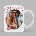 White ceramic mug featuring a romantic theme with a photo of a couple in a circular frame. The design includes pink heart-shaped confetti and a small pink heart at the bottom. Ideal for personalised gifts, Valentine's Day, or special occasions.