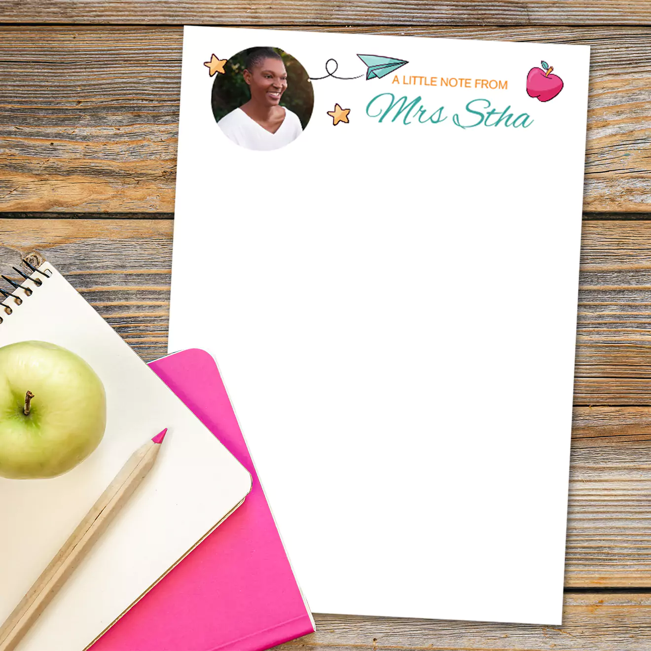 Personalised notepad with 'A Little Note from Mrs Stha' printed at the top, featuring a photo of a teacher, a paper plane, and an apple illustration. The notepad is placed on a wooden surface alongside a green apple, a pink notebook, and a pencil. Ideal for teacher gifts and personalised stationery.