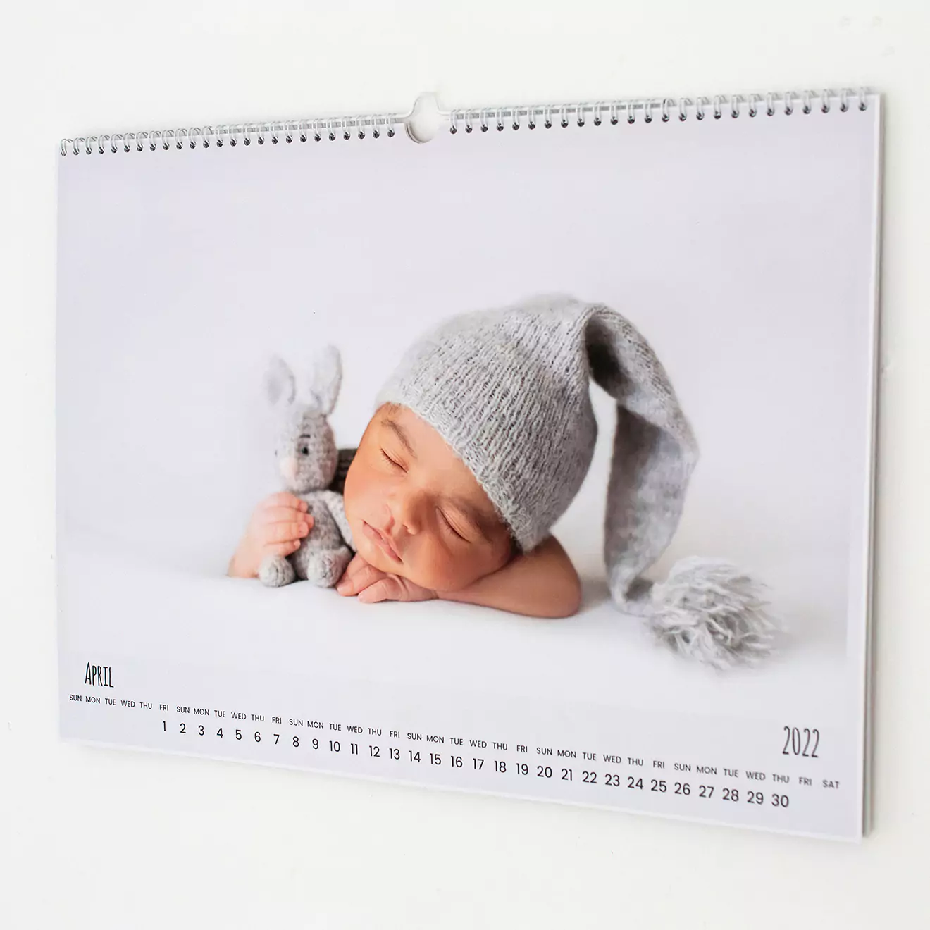 A wall calendar featuring a high-quality photograph of a sleeping baby wearing a grey knitted hat and cuddling a small grey stuffed animal. The calendar is open to April 2022, with dates clearly visible. Ideal for baby shower gift ideas or personalised gifts in South Africa.