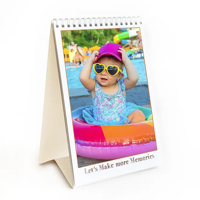 Personalised desk calendar featuring a photo of a baby in a colourful inflatable pool, wearing heart-shaped sunglasses and a pink hat. The calendar has a spiral binding and a stand, with the text 'Let's Make more Memories' at the bottom. Ideal for gifts and keepsakes.