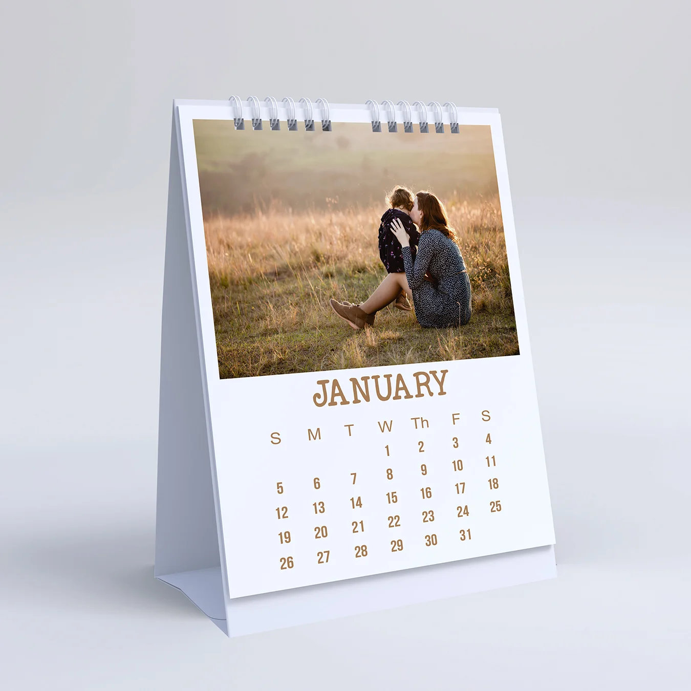 A personalised desk calendar featuring a photo of two people sitting in a field during sunset. The calendar is open to January, with the month and days clearly printed in a stylish font. The calendar has a white base and spiral binding, making it a practical and thoughtful gift idea for various occasions such as Christmas, birthdays, or Valentine's Day.