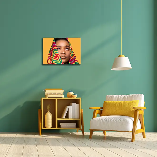 Modern living room with a green wall featuring a colourful canvas print of a person wrapped in a vibrant, patterned fabric. The room includes a wooden armchair with a yellow cushion, a matching wooden shelf with books and decorative items, and a white pendant light hanging from the ceiling. Ideal for personalised gifts in South Africa, photo to canvas, and home decor.