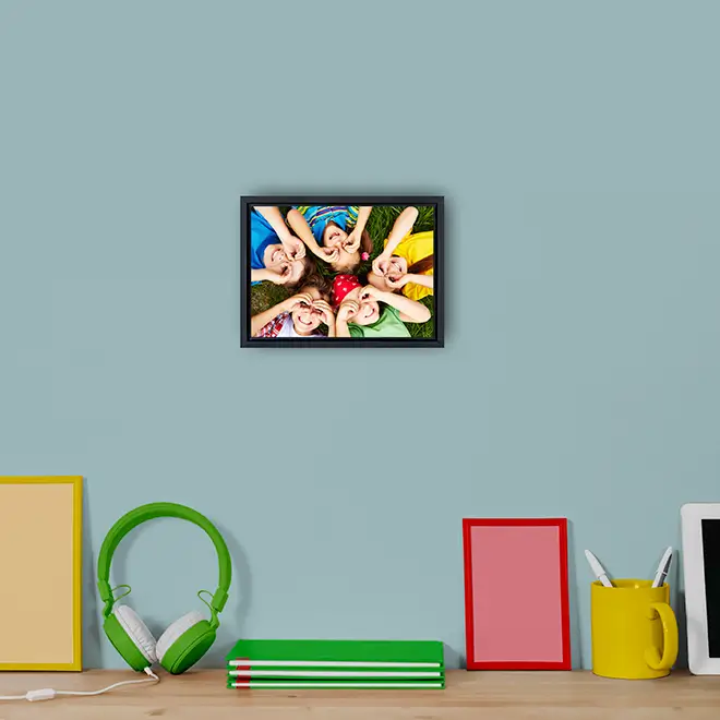 A framed photo canvas featuring a colourful image of people lying in a circle, displayed on a light blue wall. The canvas is part of a modern desk setup with green headphones, a stack of books, and various colourful desk accessories. Ideal for personalised gifts in South Africa.