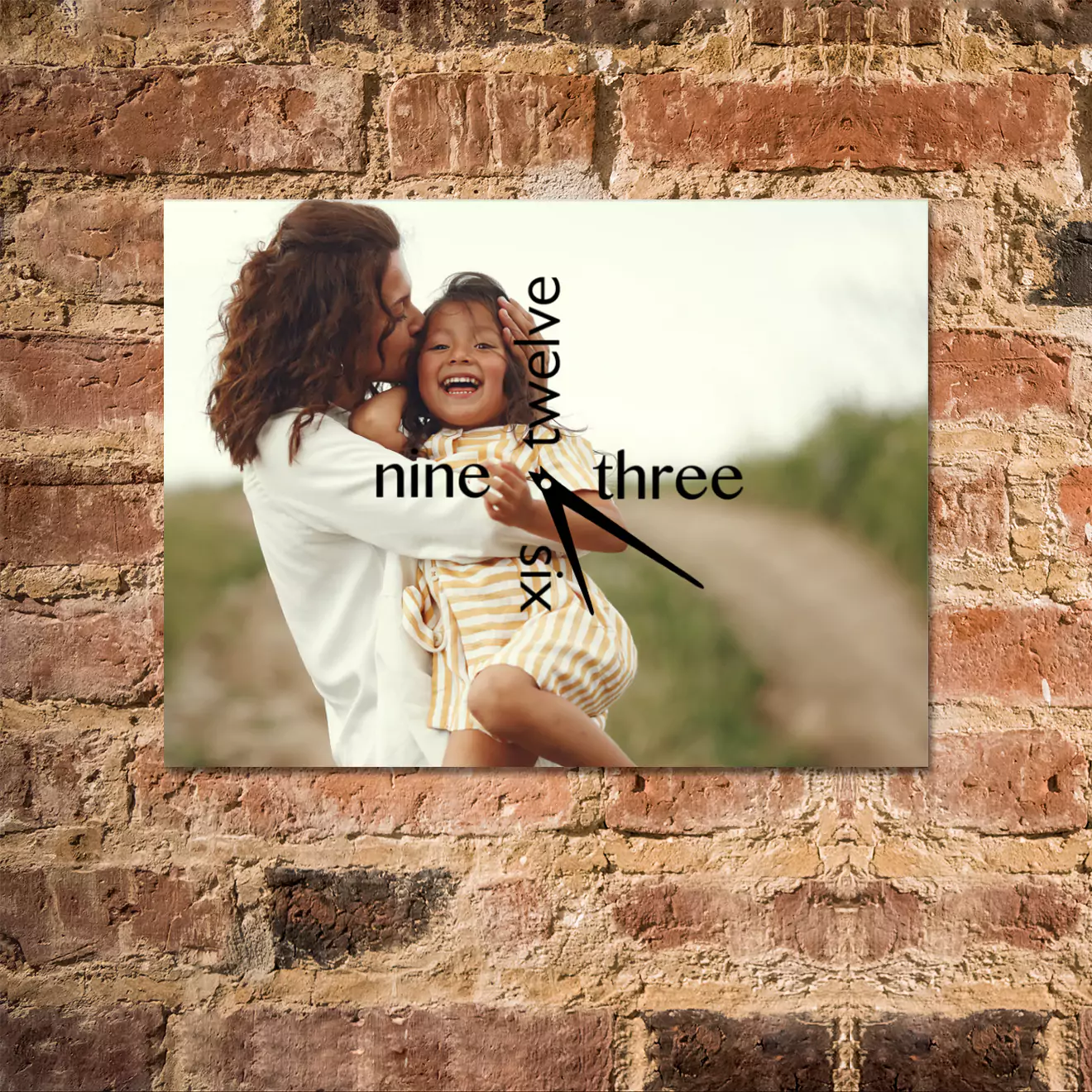 Personalised wall clock featuring a photo of a woman kissing a smiling child, set against a rustic brick wall. The clock has black hands and numbers in a modern font, with "nine," "twelve," "three," and "six" spelled out. Ideal for gifts for her, Christmas gifts, or Valentine's Day gift ideas.