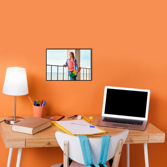 A stylish A3 canvas print featuring a vibrant photograph of a person standing by a railing, displayed on an orange wall above a wooden desk. The desk is equipped with a lamp, laptop, books, and stationery, creating a cosy and organised workspace. Ideal for personalised gifts and home decor.