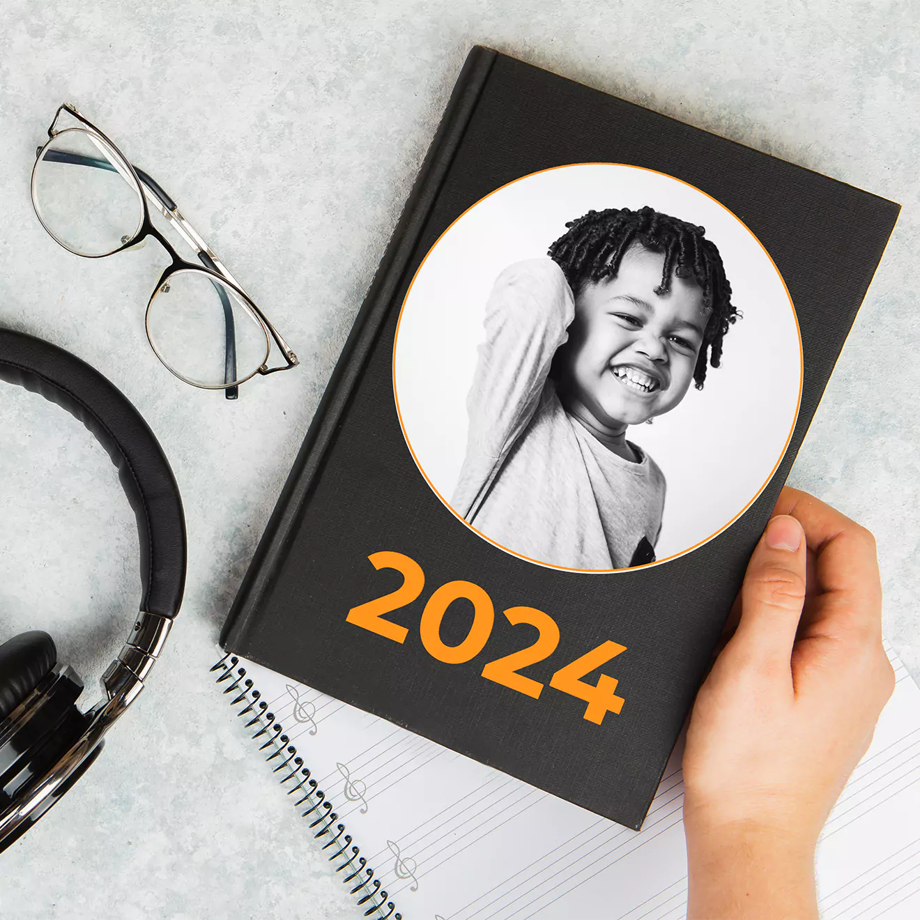 A black 2024 diary with a personalised cover featuring a smiling child in a circular frame. The diary is held by a hand and is placed on a grey surface alongside a pair of glasses, headphones, and a spiral notebook. Ideal as a personalised gift for various occasions.