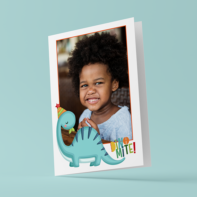 A personalised greeting card featuring a smiling child with curly hair, framed by a white border. The card has a playful dinosaur illustration wearing a party hat and the text "DINO-MITE!" in colourful letters. Ideal for baby shower gift ideas or children's birthday celebrations.