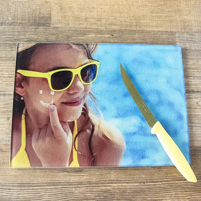 Personalised glass cutting board featuring a vibrant image of a girl wearing yellow sunglasses and a yellow swimsuit, smiling with sunscreen on her face. The cutting board is placed on a wooden surface with a yellow-handled knife beside it. Ideal for unique kitchen decor or as a thoughtful gift.