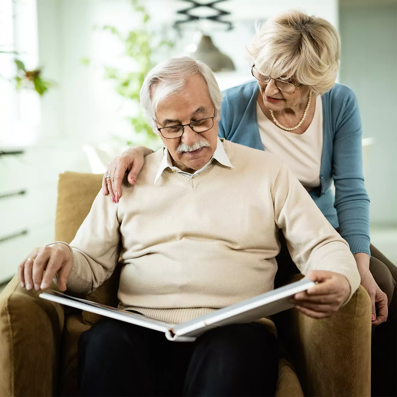 Elderly couple sitting together, looking at a large photo album. The man is seated in a beige armchair, while the woman stands beside him, resting her hand on his shoulder. The setting is a cosy, well-lit living room.