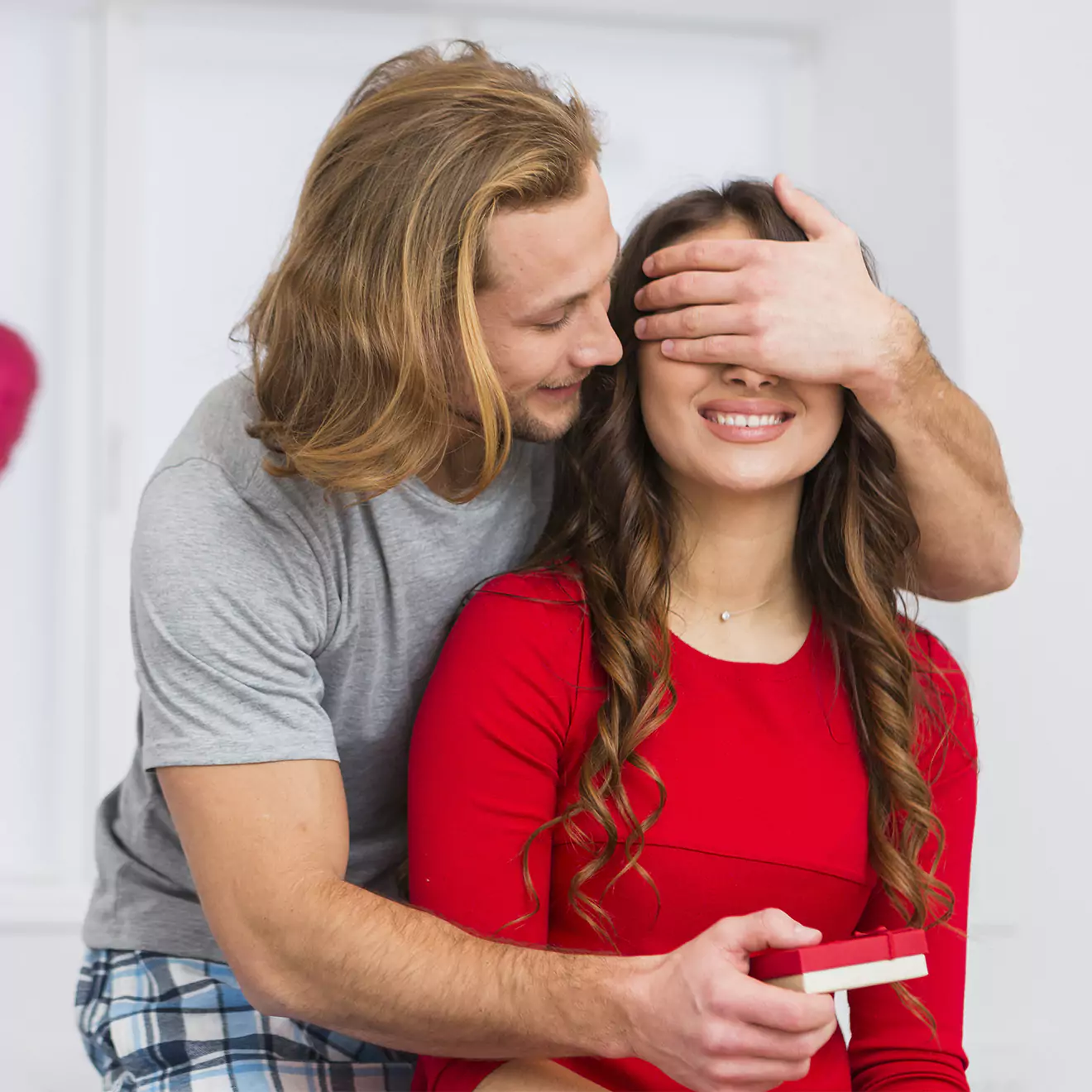 Image of a man playfully covering a woman's eyes while presenting her with a gift, depicting a sweet and romantic Valentine's Day surprise, ideal for showcasing thoughtful gift-giving.