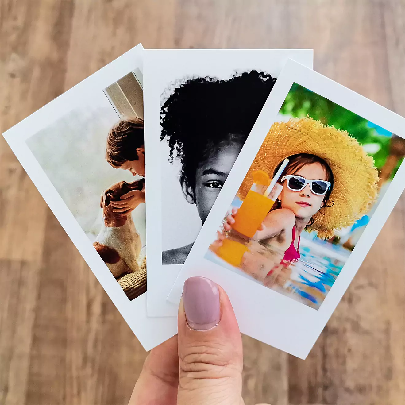 A hand holding three small, printed photos. The first photo shows a person with a dog, the second is a black and white portrait of a person, and the third features a child wearing sunglasses and a sun hat, holding a drink by the pool. Ideal for personalised gifts in South Africa or photo albums.