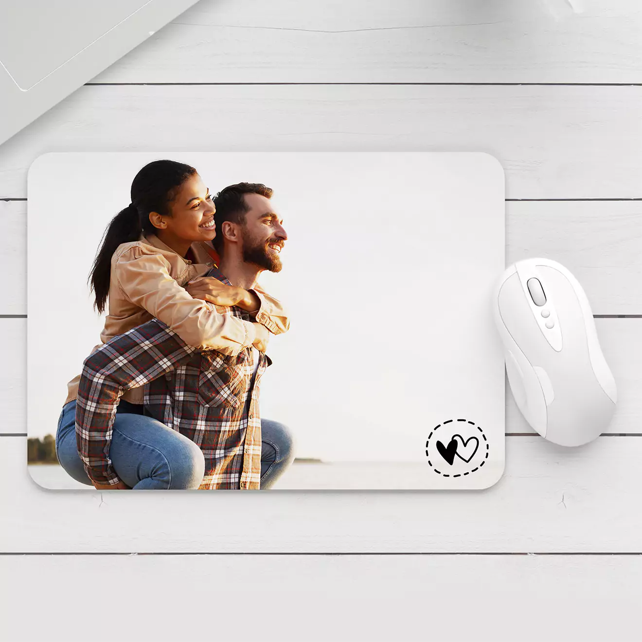 Custom mouse pad featuring a photo of a smiling couple, with the woman giving the man a piggyback ride. The mouse pad has a small heart design in the corner and is placed next to a white wireless mouse on a white wooden surface.