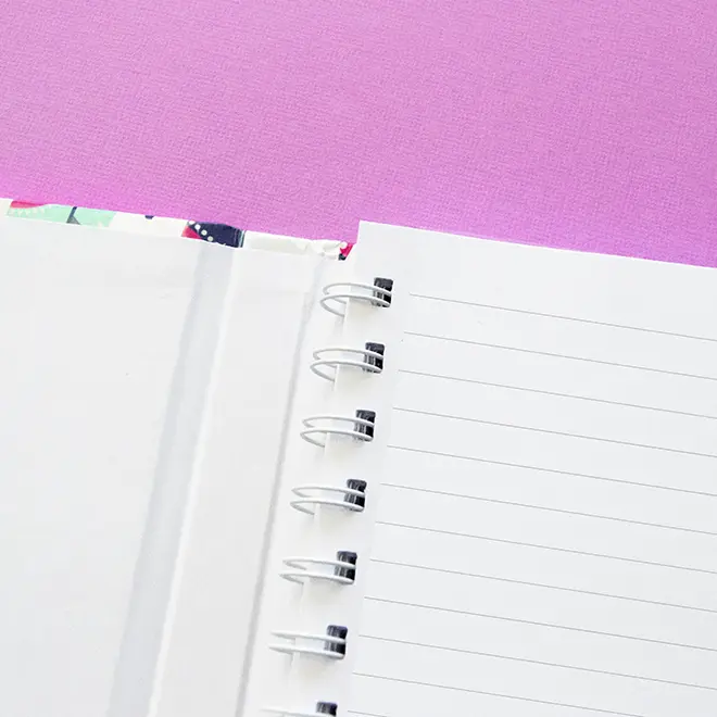 Close-up of an open spiral-bound notebook with lined pages on a vibrant pink background. The notebook features a sturdy white cover and metal rings. Ideal for note-taking, journaling, or as a personalised gift. Suitable for various occasions such as birthdays or Christmas.