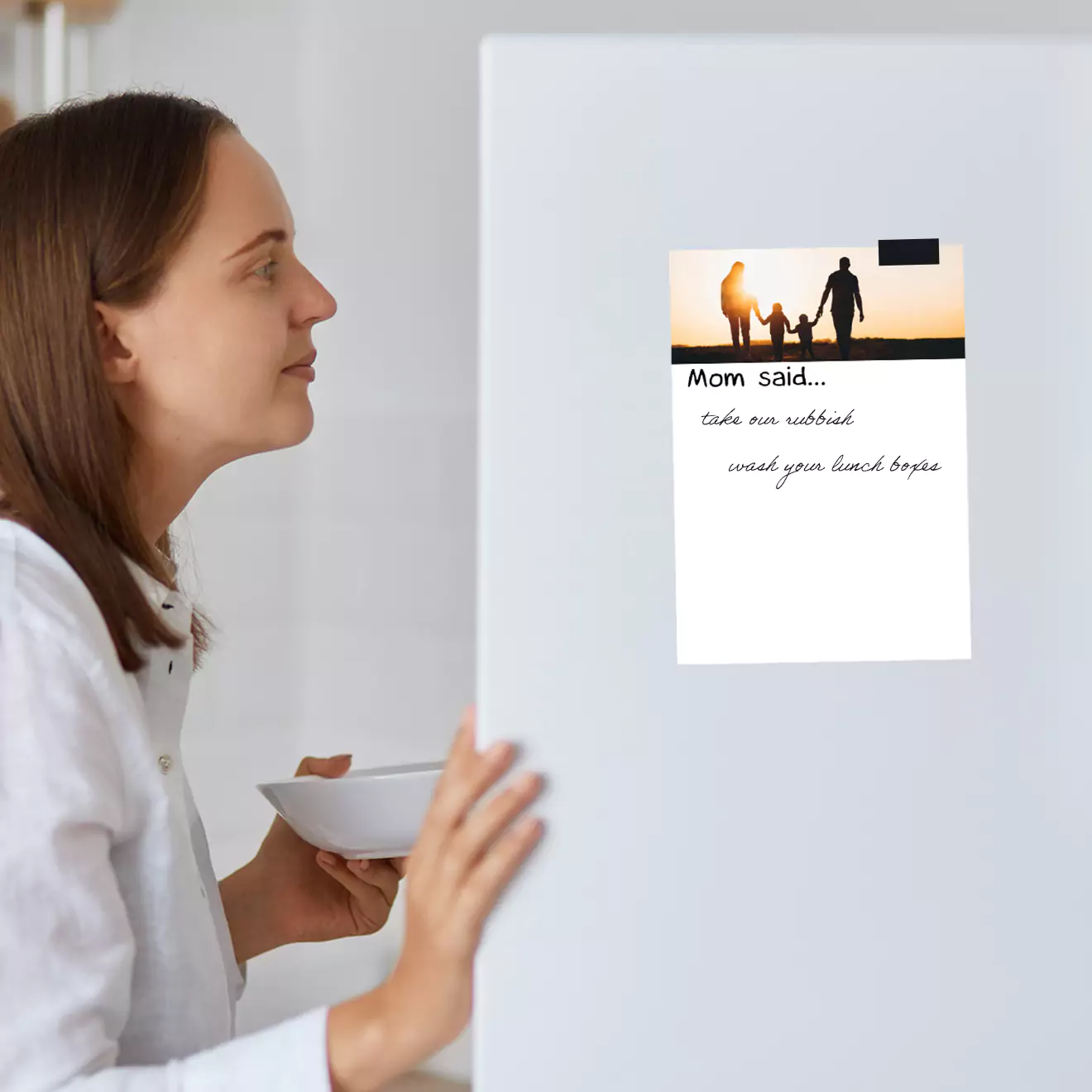 Woman looking at a personalised magnetic photo print on a refrigerator. The print features a family silhouette image at sunset and a message that reads "Mom said... take an umbrella, wash your hands before dinner." Ideal for personalised gifts in South Africa, Christmas gifts, and photo to book ideas.