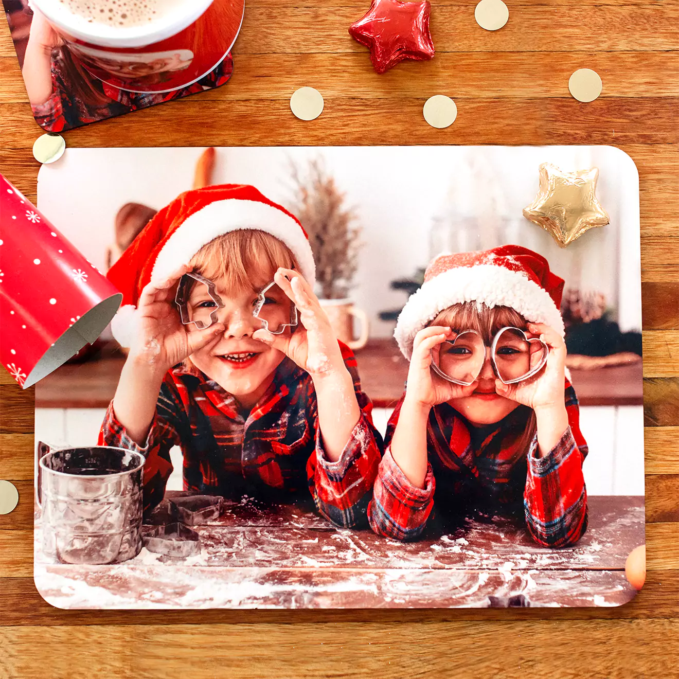 Festive photo of two children wearing Santa hats and plaid shirts, playfully holding cookie cutters over their eyes. They are surrounded by baking supplies on a wooden table, with Christmas decorations like stars and confetti scattered around. Ideal for personalised gifts in South Africa, Christmas gift ideas, and photo books.