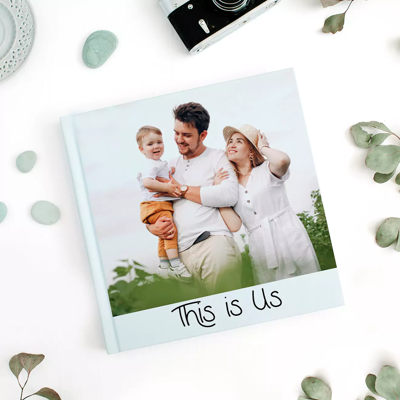 Personalised photo book titled ''This is Us'' featuring a family photo on the cover. The book has a light blue cover and is surrounded by decorative elements including a camera, leaves, and stones. Ideal for gifts for men, baby shower gift ideas, Christmas gifts, and Valentine''s Day gift ideas.