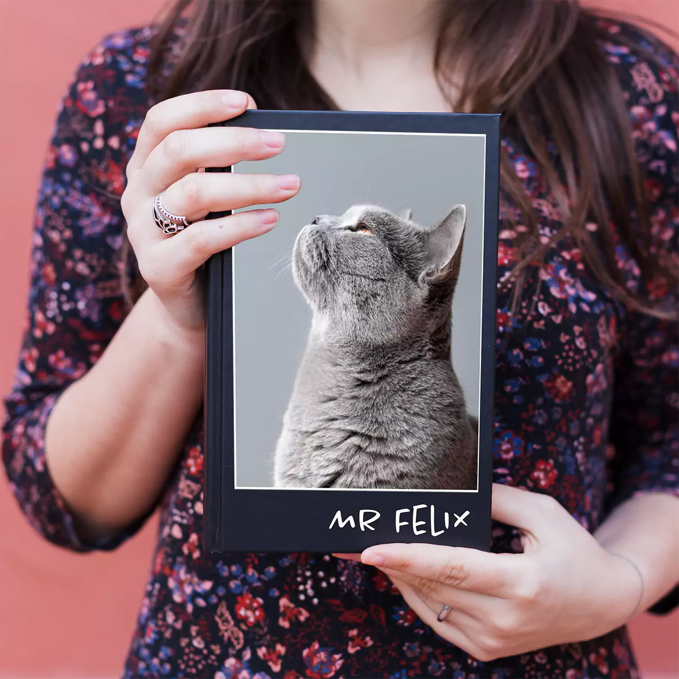 A person holding a personalised photo book with a black cover featuring a picture of a grey cat looking upwards and the text "Mr Felix" at the bottom. The person is wearing a floral-patterned top. Ideal for gifts for cat lovers, personalised gifts in South Africa, and photo albums.