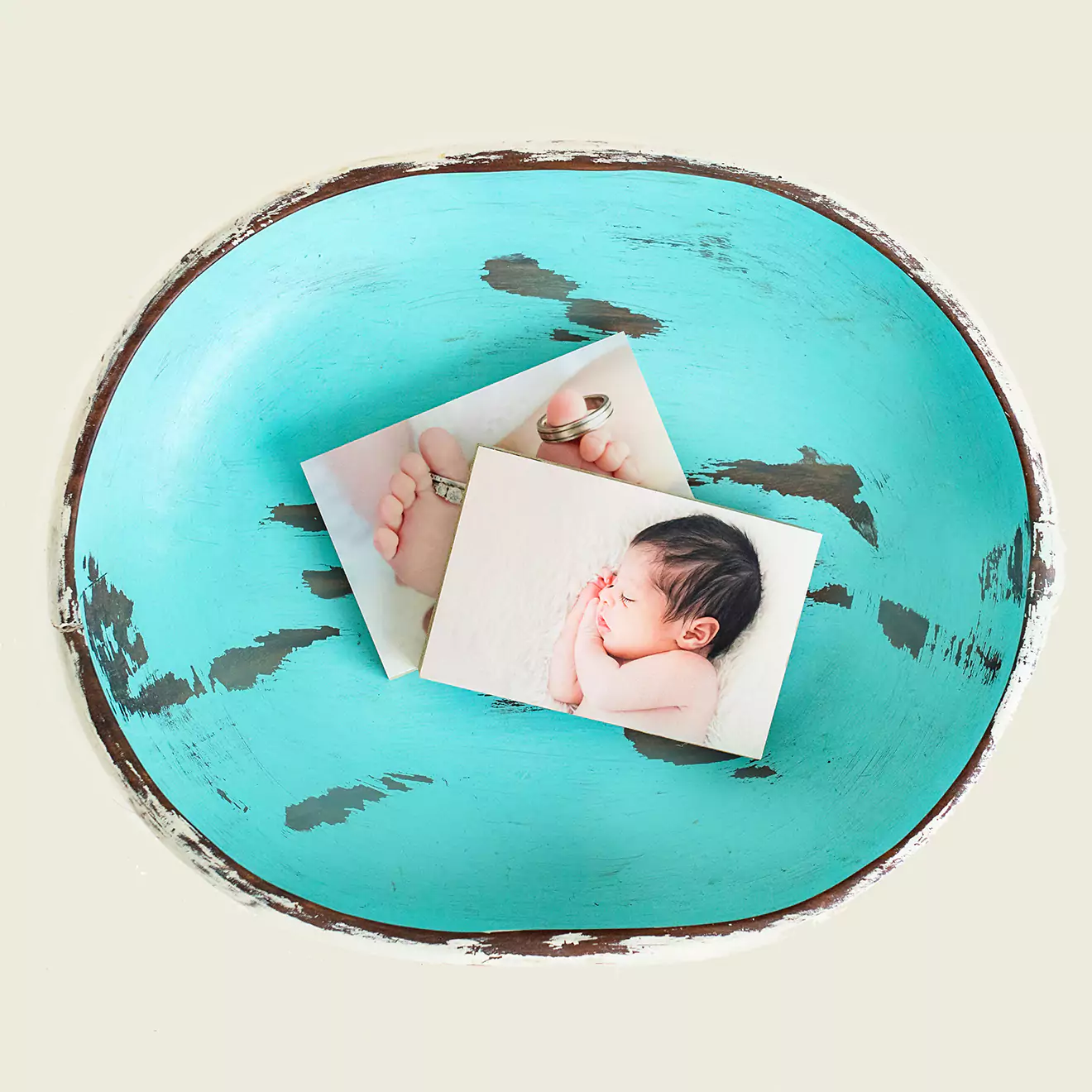 Two printed photos of a baby placed on a rustic, turquoise, distressed wooden bowl. The photos feature close-up shots of a newborn's face and tiny feet.