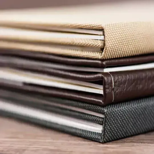 Stack of three elegant photo albums with textured covers in beige, brown, and dark grey. The albums are neatly arranged on a wooden surface, showcasing their high-quality binding and durable materials. Ideal for personalised gifts, photo books, or special occasions.