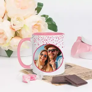 Valentine-themed custom photo mug with pink handle and rim, featuring a couple's picture. The mug is placed on a table with chocolates, candies, and a bouquet of flowers in the background.