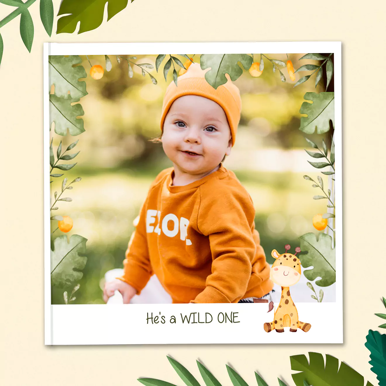 Children's photobook cover featuring a baby in an orange outfit with a wooden frame surrounded by leaves, a lion, and an elephant illustration. Text reads 'He's a WILD ONE'.