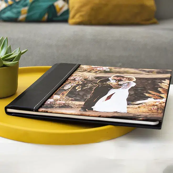 A stylish photo book with a black spine and a custom cover featuring a wedding photo, placed on a yellow round tray beside a small potted succulent. The photo book is ideal for preserving cherished memories and makes a perfect gift for occasions such as weddings, anniversaries, or Valentine's Day.