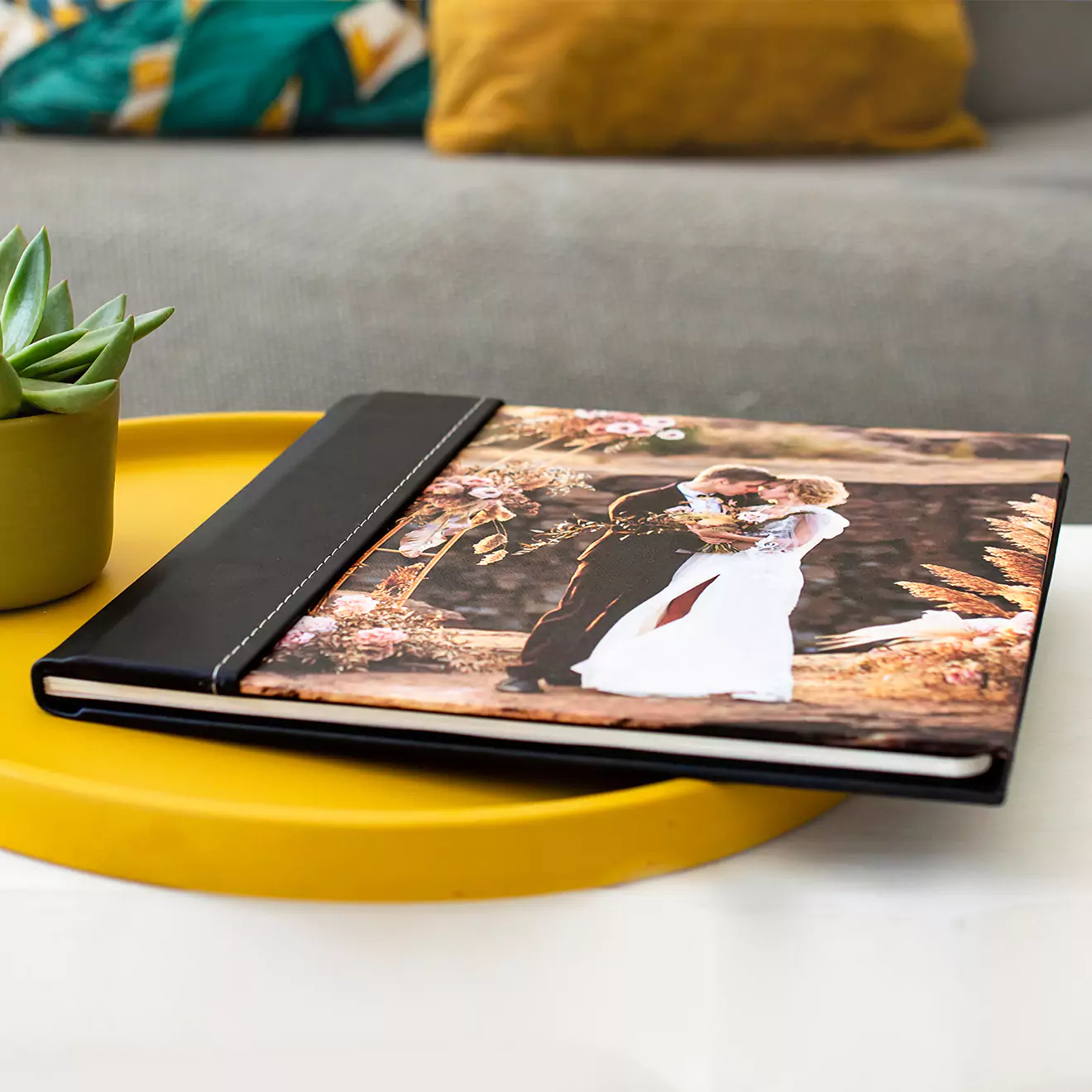 Elegant personalised photo book with a black spine and a cover featuring a couple in wedding attire, placed on a yellow tray next to a small green potted plant. Ideal for wedding memories, gifts for men, baby shower gift ideas, Christmas gifts, and Valentine's Day gift ideas.