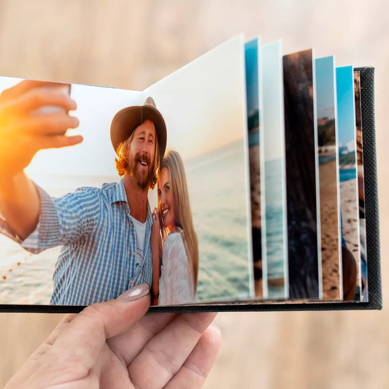 A hand holding an open photo book displaying vibrant, high-quality images of a couple enjoying a beach setting. The photo book features a durable black cover and thick pages, making it an ideal choice for personalised gifts in South Africa, photo albums, or special occasion keepsakes.