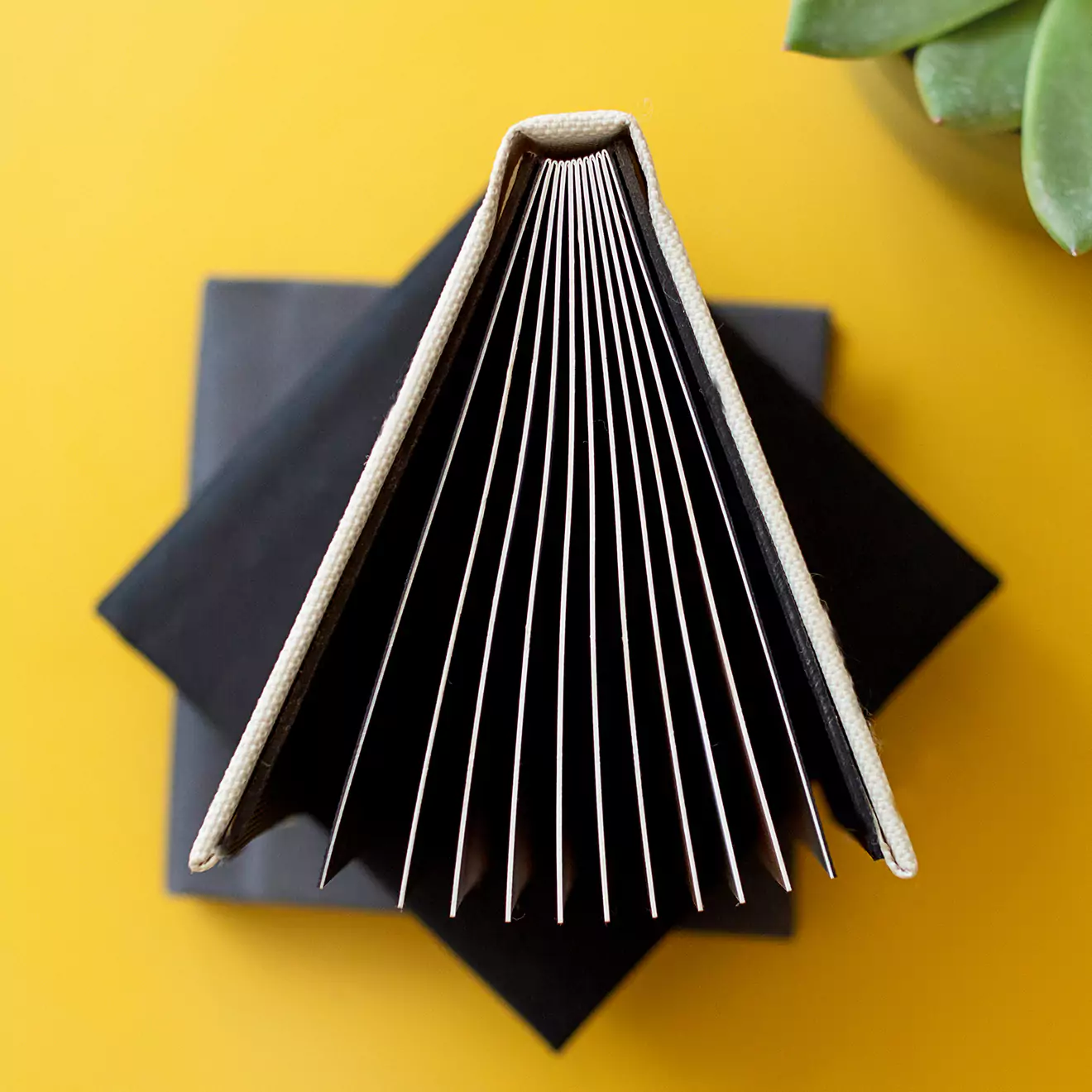 A top-down view of an open photo book with a beige cover, displaying its pages fanned out. The book is placed on a black square surface against a bright yellow background. Ideal for personalised gifts, photo albums, and special occasions such as Christmas or Valentine's Day.