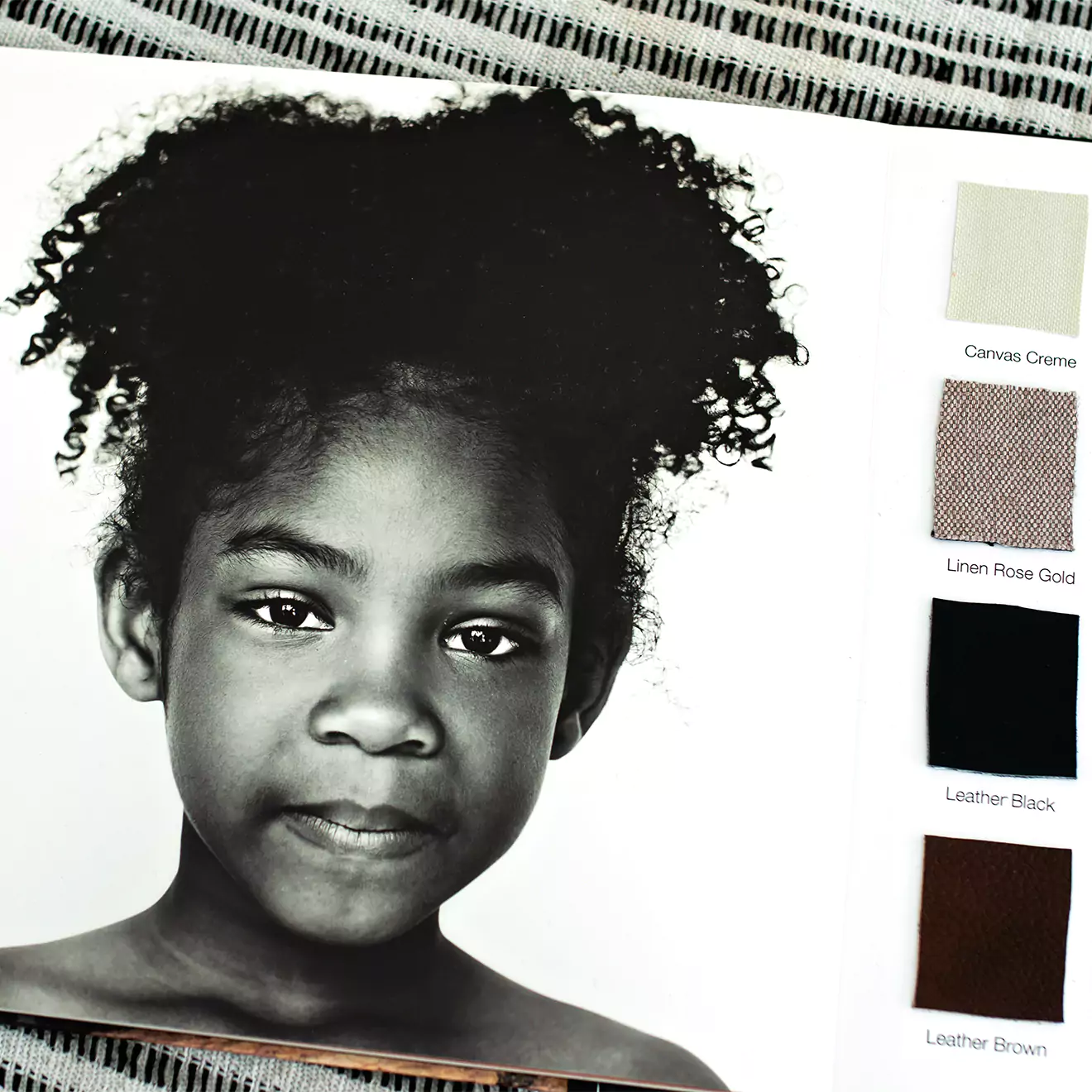 Open photo book displaying a black and white portrait of a young girl with curly hair. Beside the photo are fabric swatches in four colours: Canvas Creme, Linen Rose Gold, Leather Black, and Leather Brown. The book is placed on a textured surface. Ideal for personalised gifts in South Africa.