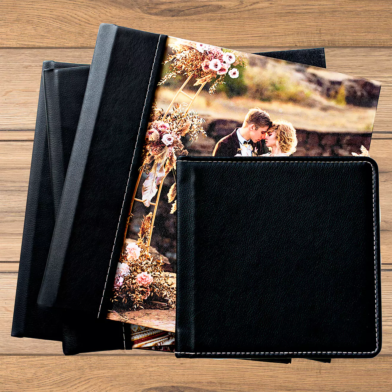 A collection of elegant black photo albums with a leather-like finish, displayed on a wooden surface. One album is open, showcasing a wedding photograph with a couple embracing amidst a floral backdrop. Ideal for preserving cherished memories, these albums make thoughtful gifts for various occasions such as weddings, anniversaries, and birthdays.