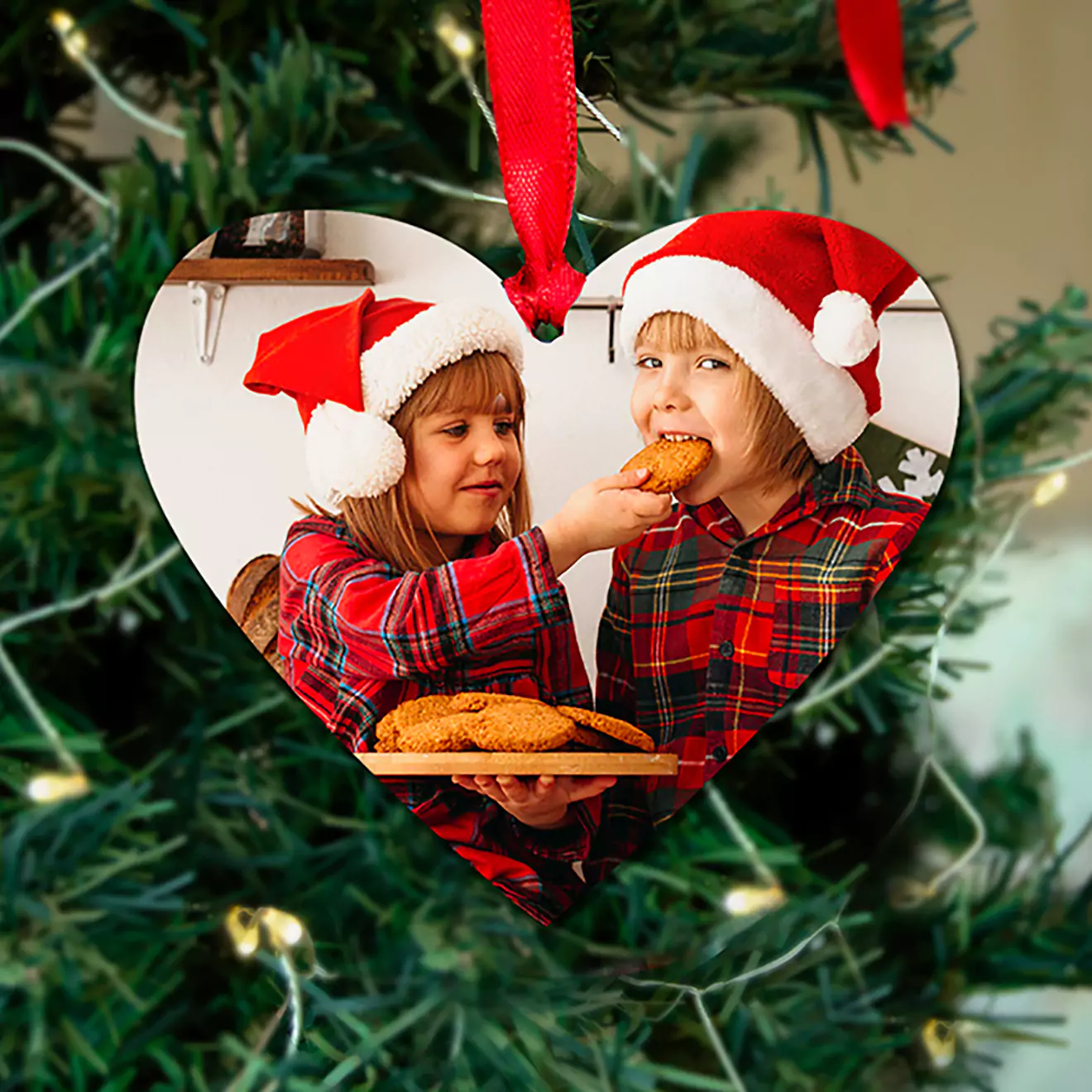 Heart-shaped Christmas tree ornament featuring a photo of two children in festive attire, wearing red Santa hats and plaid shirts, sharing cookies. The ornament is hung with a red ribbon and is set against a backdrop of a decorated Christmas tree. Ideal for personalised Christmas gifts.