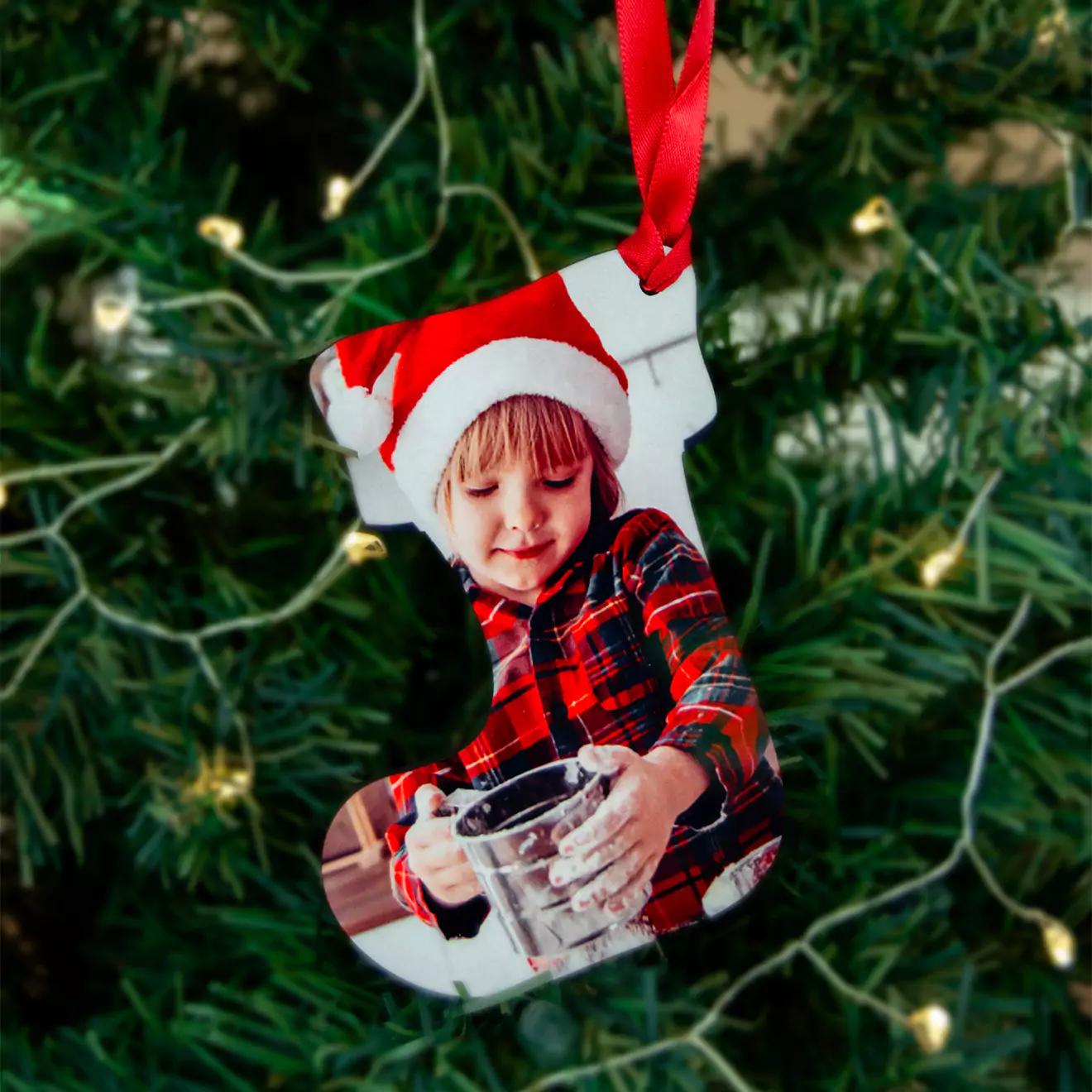 Christmas tree decoration featuring a personalised stocking-shaped ornament with a photo of a child wearing a Santa hat and plaid shirt. The ornament hangs from a red ribbon and is set against a backdrop of green Christmas tree branches adorned with fairy lights. Ideal for Christmas gift ideas and personalised gifts in South Africa.