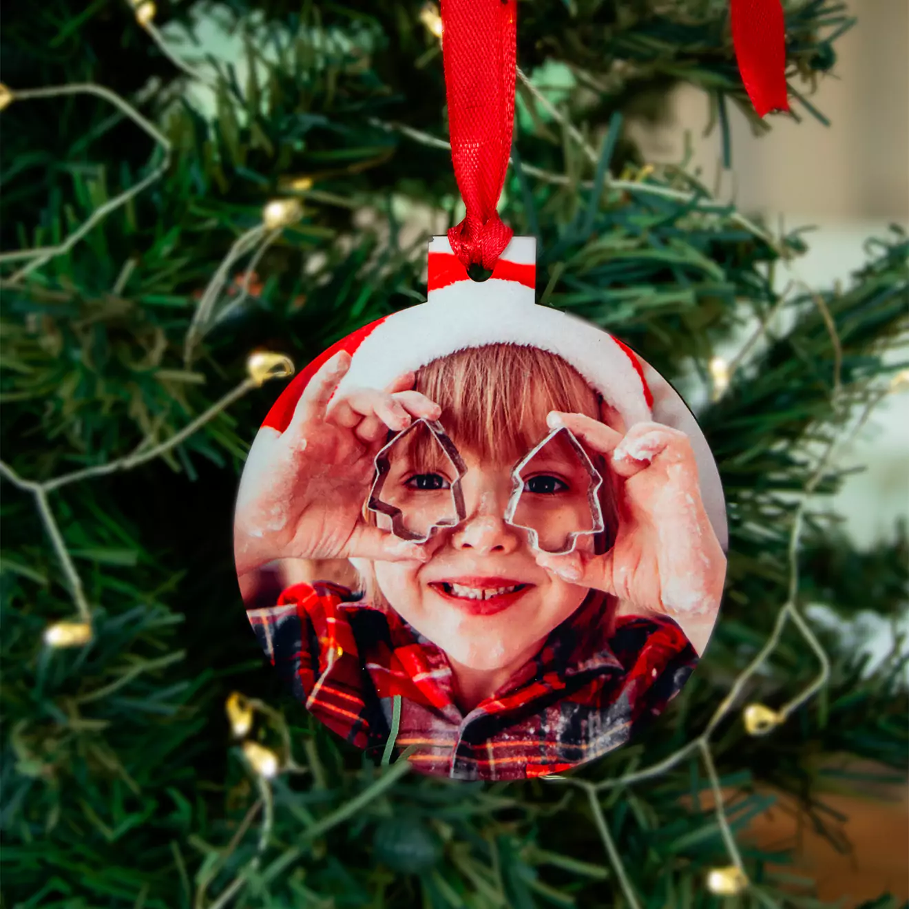 A personalised Christmas tree decoration featuring a circular ornament with a photo of a smiling child wearing a Santa hat and holding cookie cutters shaped like Christmas trees. The ornament is hanging on a green Christmas tree with fairy lights and a red ribbon. Ideal for Christmas gift ideas and personalised gifts in South Africa.