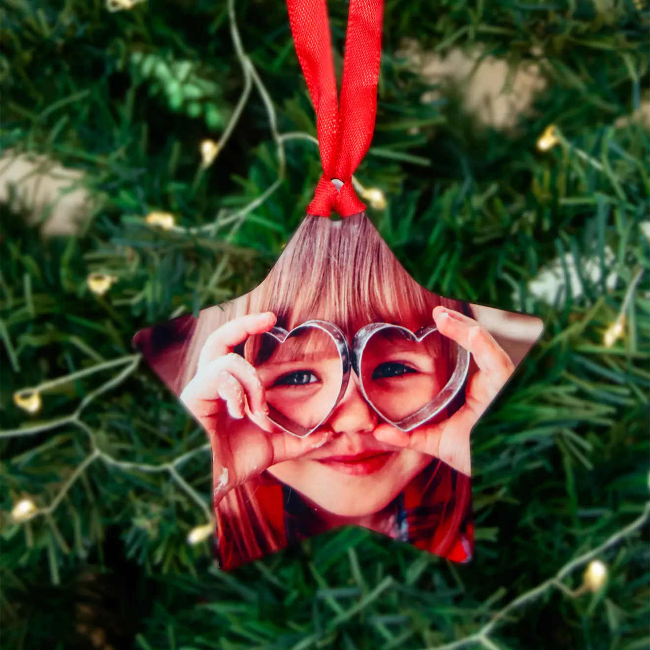 Star-shaped Christmas tree ornament featuring a personalised photo of a child with heart-shaped glasses, hanging from a red ribbon. The ornament is set against a background of green Christmas tree branches with small, warm white lights. Ideal for personalised gifts and Christmas decorations.