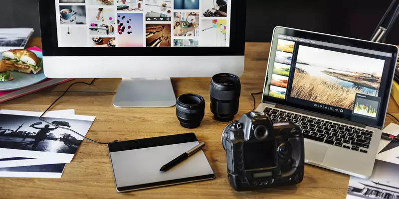 Desk cluttered with a camera in front of two PCs, a tablet, and scattered photos, capturing a creative workspace